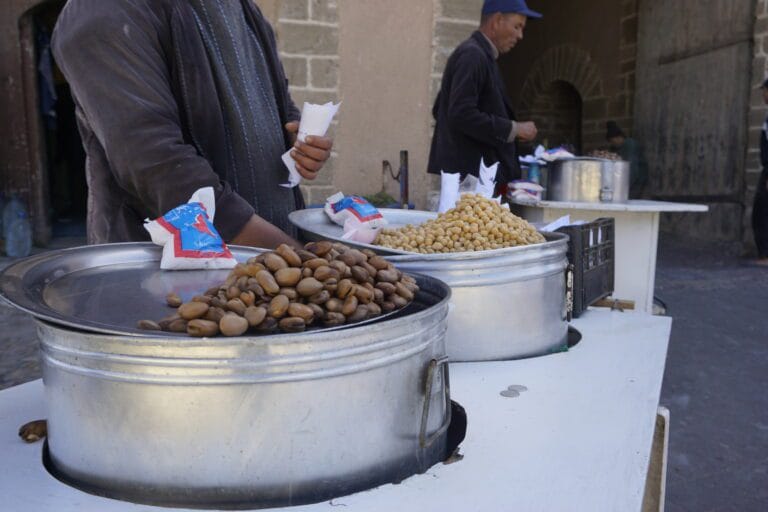 A man stands by a table of Moroccan street food, showcasing delicious beans and chickpeas.